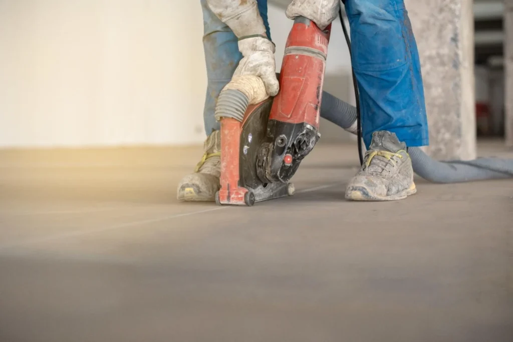 A construction worker using a red power saw with a vacuum attachment to cut a straight expansion joint into a concrete floor.